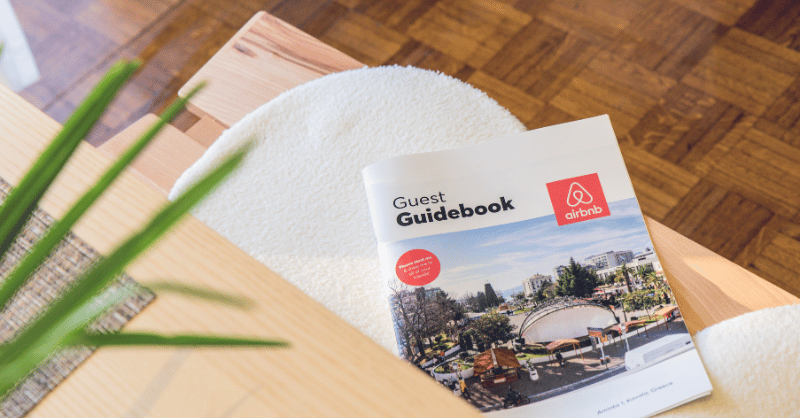 A guest guidebook with an Airbnb logo lies open on a light-colored table, perfect for those seeking short-term rental advice. The cover showcases a cityscape with trees and people. Nearby, the corner of a textured cushion and a blurred green plant enhance its optimal visibility.