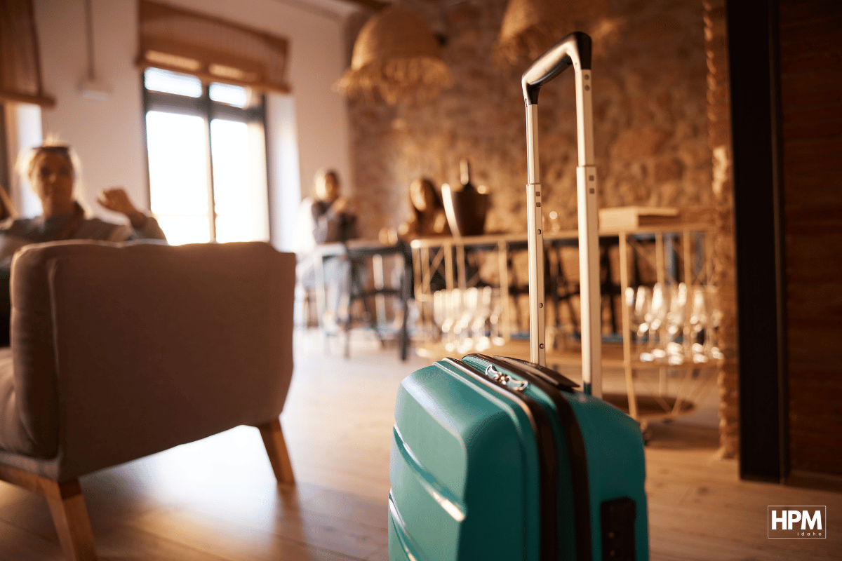 A turquoise suitcase stands in focus in the foreground, highlighting optimal visibility for your short-term rental. In the background, people relax on couches and chairs in an open-plan room with stone walls and wooden decor, creating a cozy atmosphere.