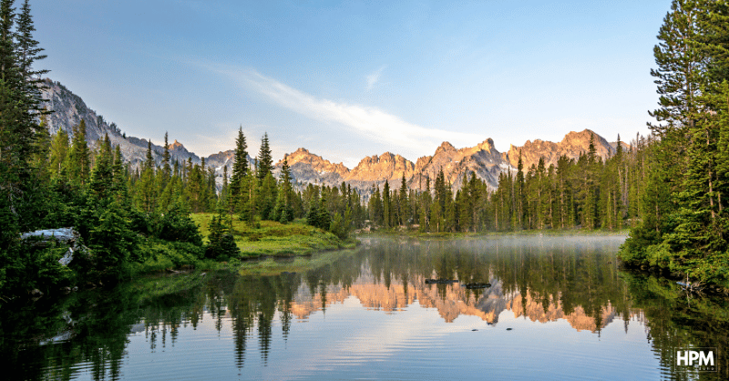 A serene lake reflects the rocky mountain peaks and evergreen trees, offering an ideal view for a short-term rental. The early morning light casts a gentle glow on the landscape, while mist rises from the waters surface, ensuring optimal visibility on any guest’s list of must-see sights.