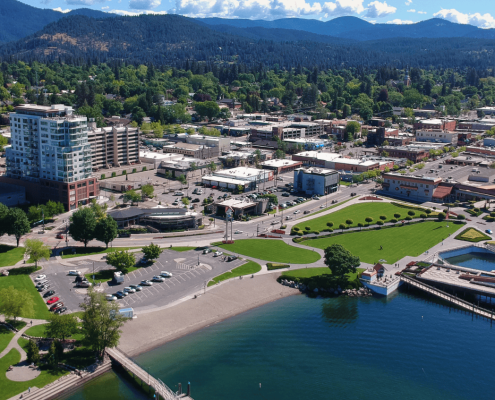 Aerial view of downtown Coeur d'Alene, Idaho, showcasing real estate opportunities with green park spaces, a parking lot, and nearby buildings. Surrounding mountains, trees, and a clear blue sky complete this picturesque setting.