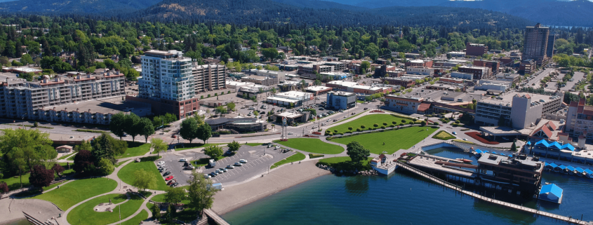 Aerial view of downtown Coeur d'Alene, Idaho, showcasing real estate opportunities with green park spaces, a parking lot, and nearby buildings. Surrounding mountains, trees, and a clear blue sky complete this picturesque setting.