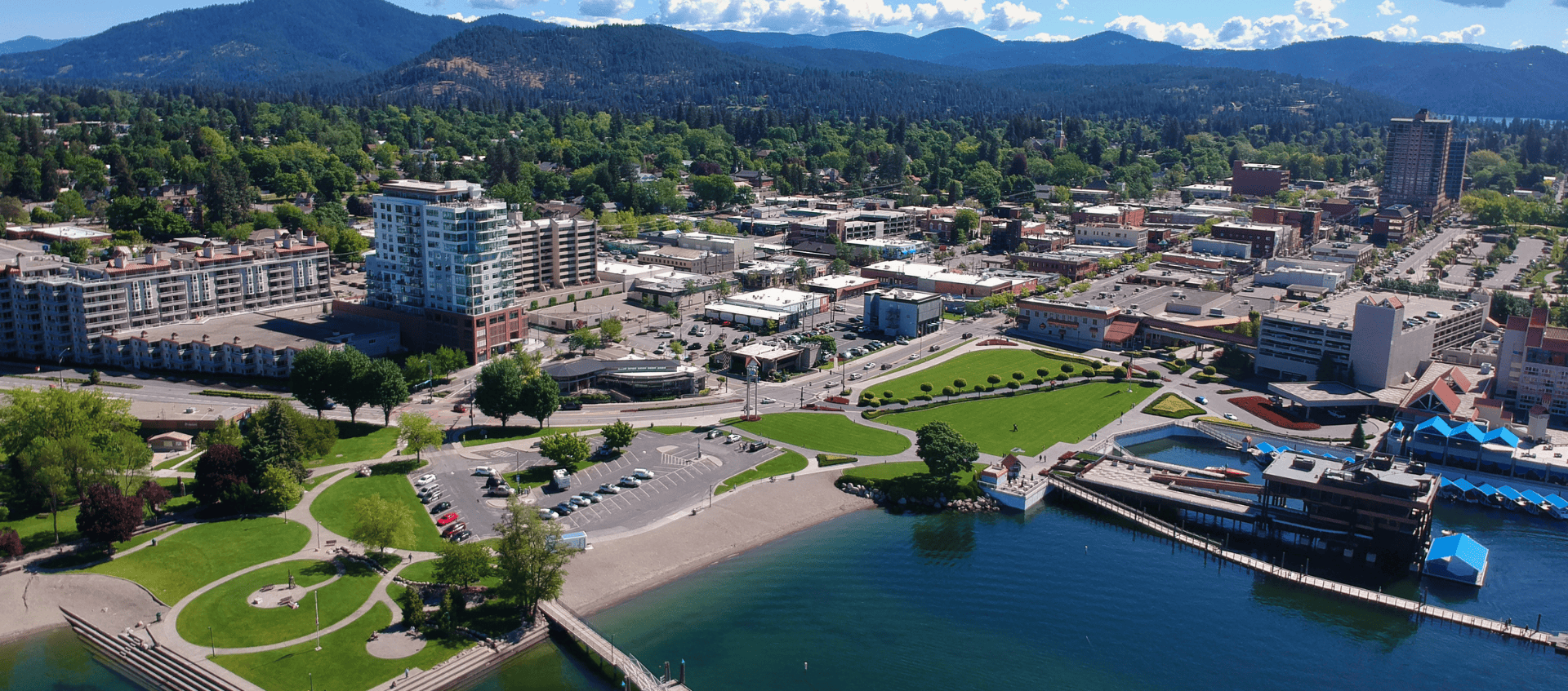 Aerial view of downtown Coeur d'Alene, Idaho, showcasing real estate opportunities with green park spaces, a parking lot, and nearby buildings. Surrounding mountains, trees, and a clear blue sky complete this picturesque setting.