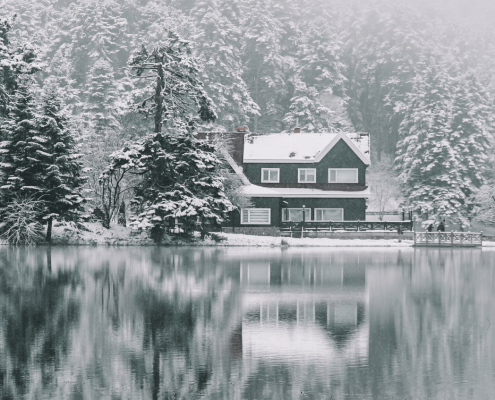 A cozy cabin surrounded by snow-covered pine trees sits at the edge of a still, reflective lake in Coeur d'Alene. Fog blankets the forest, creating a serene and tranquil winter scene that's perfect for Cozy Lakefront Vacation Rentals.