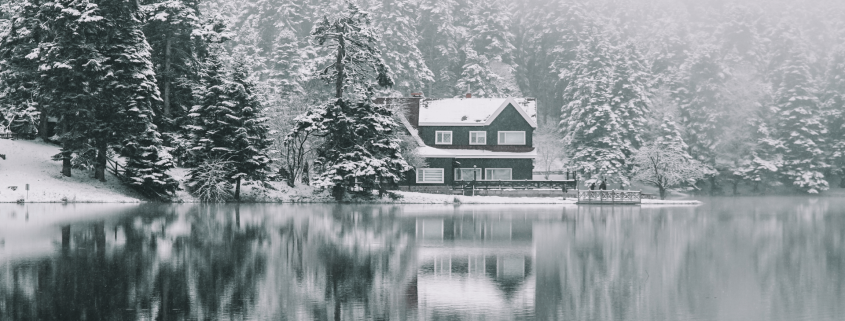 A cozy cabin surrounded by snow-covered pine trees sits at the edge of a still, reflective lake in Coeur d'Alene. Fog blankets the forest, creating a serene and tranquil winter scene that's perfect for Cozy Lakefront Vacation Rentals.