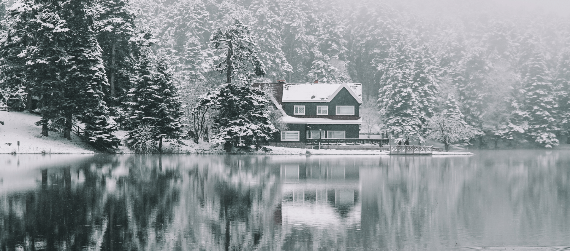 A cozy cabin surrounded by snow-covered pine trees sits at the edge of a still, reflective lake in Coeur d'Alene. Fog blankets the forest, creating a serene and tranquil winter scene that's perfect for Cozy Lakefront Vacation Rentals.