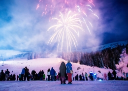 A lively crowd, bundled in winter clothing, watches colorful fireworks illuminate the night sky over Sandpoint's snow-covered landscape, where trees and a ski slope create a perfect backdrop for one of the town's cherished winter festivals.