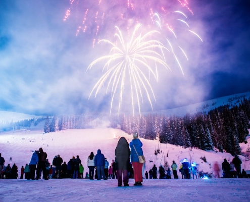 A lively crowd, bundled in winter clothing, watches colorful fireworks illuminate the night sky over Sandpoint's snow-covered landscape, where trees and a ski slope create a perfect backdrop for one of the town's cherished winter festivals.