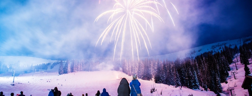 A lively crowd, bundled in winter clothing, watches colorful fireworks illuminate the night sky over Sandpoint's snow-covered landscape, where trees and a ski slope create a perfect backdrop for one of the town's cherished winter festivals.