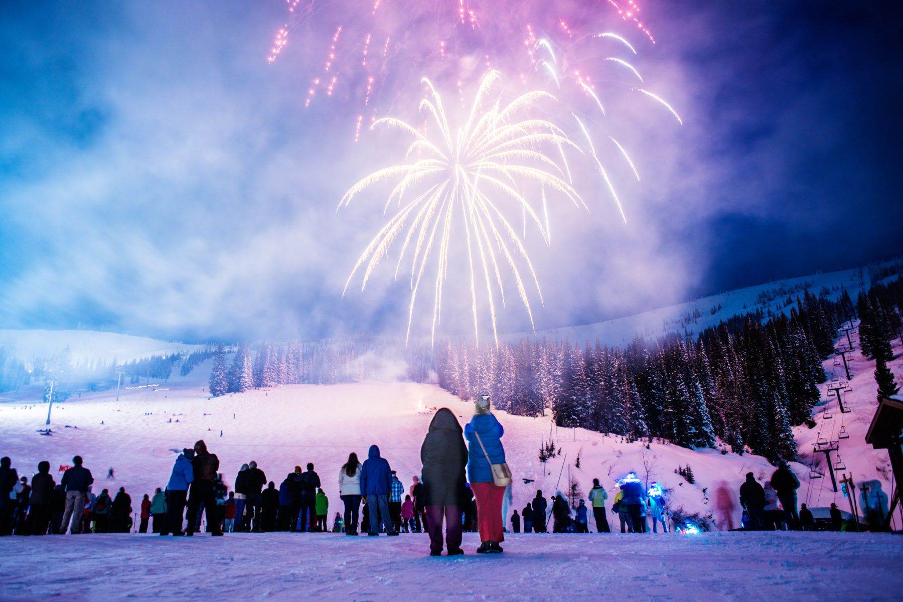 A lively crowd, bundled in winter clothing, watches colorful fireworks illuminate the night sky over Sandpoint's snow-covered landscape, where trees and a ski slope create a perfect backdrop for one of the town's cherished winter festivals.