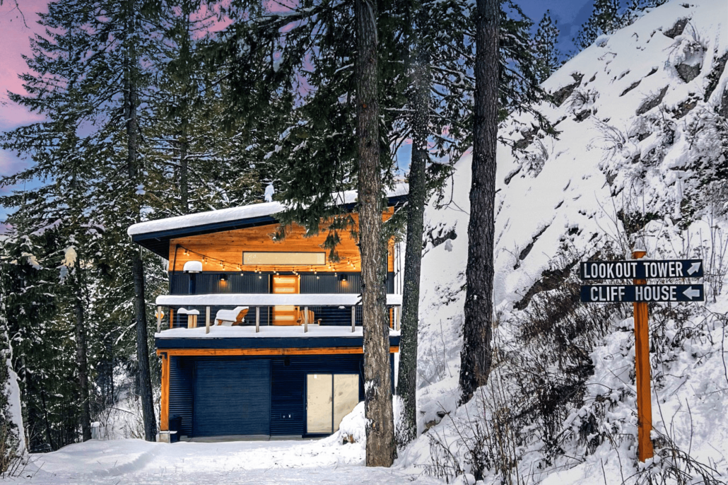 A modern wooden cabin with large glass doors is nestled among snow-covered trees and rocks at Schweitzer Mountain Resort. It features a balcony and two garage doors. A signpost nearby directs to Lookout Tower and Cliff House, as twilight skies provide a colorful backdrop for ski season adventures.