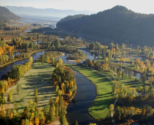 Aerial view of a scenic golf course surrounded by winding rivers and lush greenery is perfect for travel planning. The landscape, ideal for visitor resources, features autumn-colored trees and green fairways, with hills visible in the background under a clear sky.