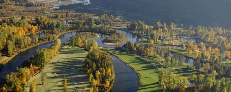 Aerial view of a scenic golf course surrounded by winding rivers and lush greenery is perfect for travel planning. The landscape, ideal for visitor resources, features autumn-colored trees and green fairways, with hills visible in the background under a clear sky.