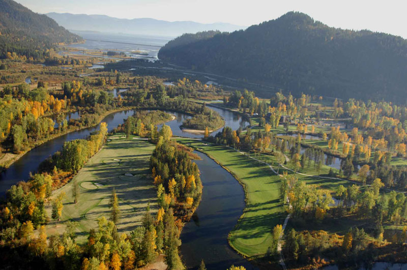 Aerial view of a scenic golf course surrounded by winding rivers and lush greenery is perfect for travel planning. The landscape, ideal for visitor resources, features autumn-colored trees and green fairways, with hills visible in the background under a clear sky.