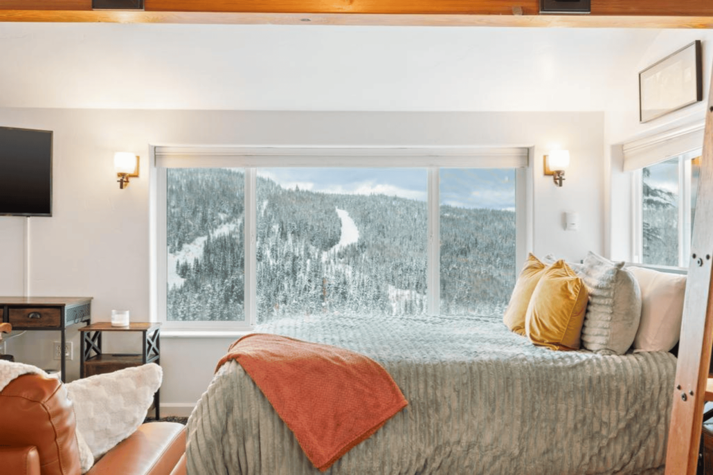 A cozy bedroom at Schweitzer Mountain Resort showcases a large window with snow-covered mountains, capturing the perfect ski season view. The room features a bed with orange and gray bedding, a leather chair, warm lighting, and a flat-screen TV mounted on the wall to the left.