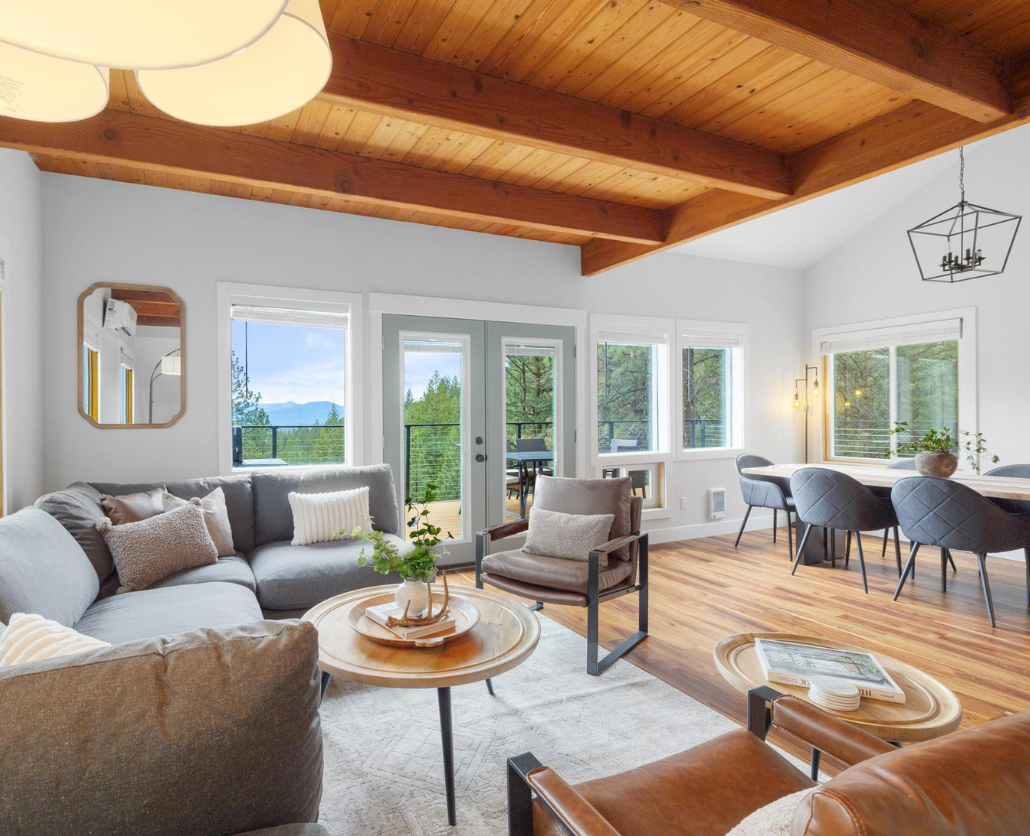 Discover a cozy, pet-friendly Sandpoint rental featuring a gray sectional sofa, wooden coffee table, and brown chair. Large windows and a glass door offer forest views. The dining area boasts a round table with modern chairs beneath wooden-beamed ceilings.