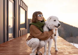 A person wearing sunglasses and a scarf kneels on a wooden deck next to a large white dog. They are outdoors near a modern cabin with a scenic view of mountains and a bright sky in the background.