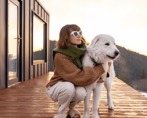 A person wearing sunglasses and a scarf kneels on a wooden deck next to a large white dog. They are outdoors near a modern cabin with a scenic view of mountains and a bright sky in the background.