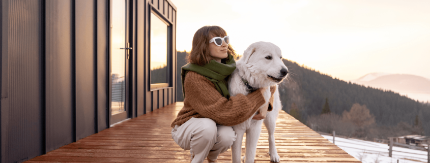 A person wearing sunglasses and a scarf kneels on a wooden deck next to a large white dog. They are outdoors near a modern cabin with a scenic view of mountains and a bright sky in the background.