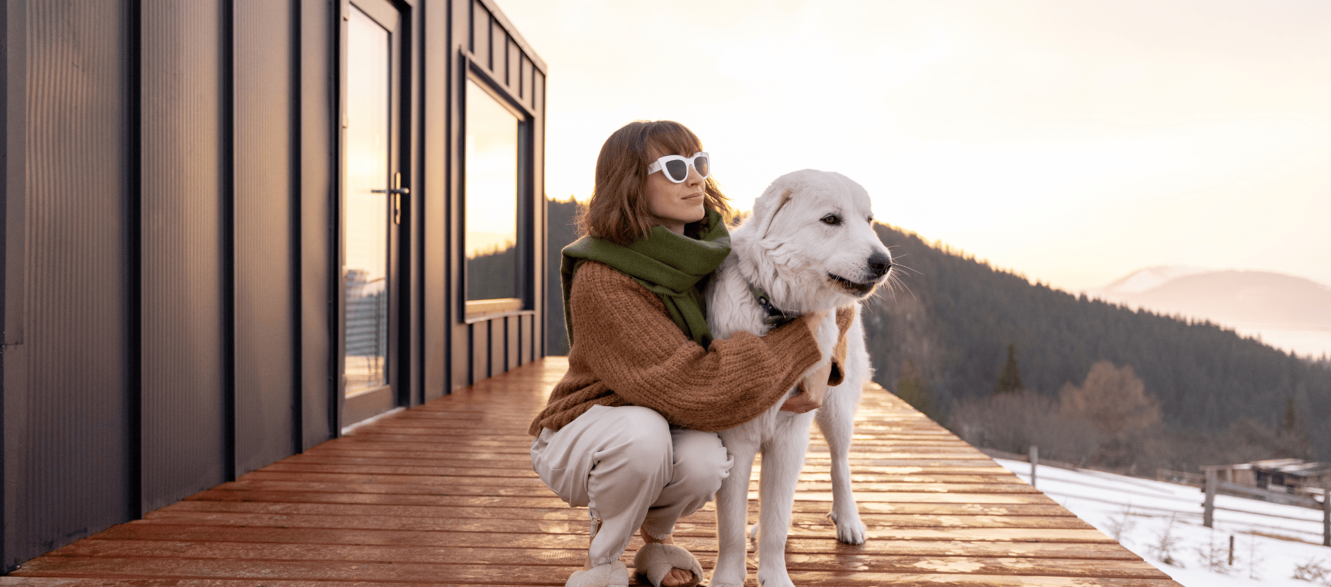 A person wearing sunglasses and a scarf kneels on a wooden deck next to a large white dog. They are outdoors near a modern cabin with a scenic view of mountains and a bright sky in the background.