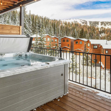 A hot tub on a wooden deck overlooks a snowy forest and mountain scene in the heart of Idaho Vacation Rentals. Cozy wooden cabins are visible in the background, surrounded by snow-covered pine trees under a partly cloudy blue sky, perfect for your vacation rentals in Coeur dAlene, Idaho.