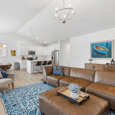 A modern living room in Idaho vacation rentals boasts a brown leather sectional sofa, blue patterned rug, and a large ottoman. A wooden dining table is visible near the airy white kitchen with bar stools. One wall features a framed picture of a boat, adding charm to this cozy getaway.