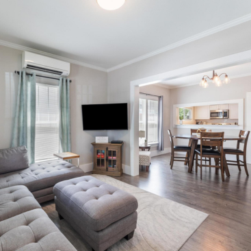 A modern living room in a chic Idaho vacation rental features a gray sectional sofa and ottoman facing a wall-mounted TV. Adjacent is a dining area with a wooden table and chairs, all aglow from large windows, with the kitchen visible in the background. Perfect for your Idaho getaway.