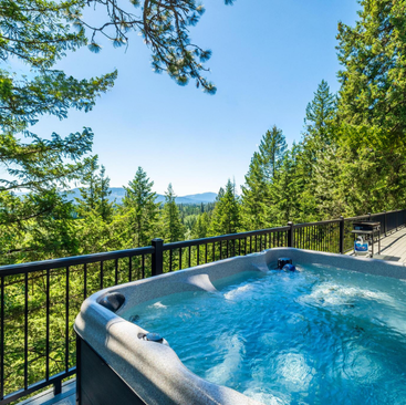 A hot tub surrounded by a railing overlooks a scenic view of lush green pine trees and distant mountains under a clear blue sky, offering the perfect retreat for those seeking Idaho vacation rentals.