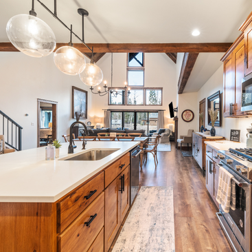 Experience the charm of Idaho Vacation Rentals with this spacious, modern kitchen featuring wooden cabinets and a large central island. Enjoy globe pendant lights, rustic beams, and hardwood floors that lead into a living area with plush seating—your ideal escape in Sandpoint or Coeur dAlene.