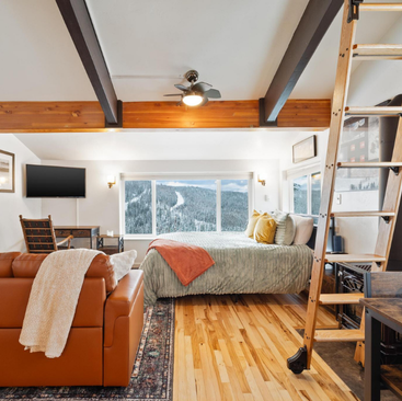 A cozy, rustic Schweitzer Vacation Rental bedroom features a large window with a scenic mountain view. It includes a wooden loft ladder, a leather sofa draped with a blanket, vibrant pillows on the bed, TV, and small work desk. Exposed ceiling beams and wooden flooring add to its charm.