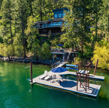 A serene lakeside scene with a dock and patio chairs under a blue umbrella invites relaxation at Hayden Lake Vacation Rentals. A house with a deck sits on a wooded hillside, surrounded by tall trees and lush greenery. The clear green water reflects the natural beauty of the area.