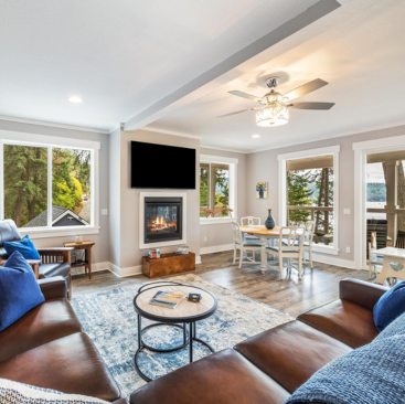 A cozy living room with brown leather sofas, a round coffee table, and a blue rug. A fireplace and TV are set against the wall. Large windows offer views of lush greenery and a lake, perfect for Idaho vacation rentals. A dining area with white chairs is visible in the background.