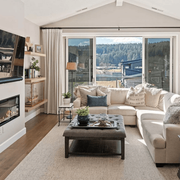 A cozy living room in one of the charming Idaho vacation rentals, featuring a beige sectional sofa, a large window with a scenic view, and a wall-mounted fireplace. Enjoy modern amenities like a flat-screen TV, potted plants, and decorative shelves that enhance the bright, inviting space.