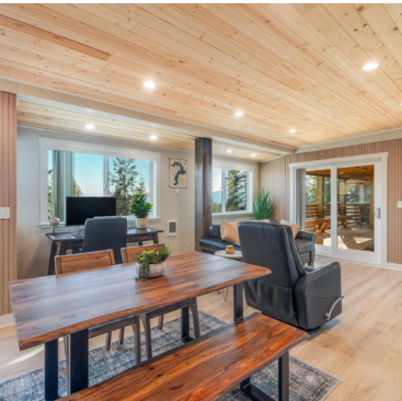 Experience a modern Sandpoint vacation rental featuring an open-concept living space with wooden ceilings and floors. This cozy retreat includes a dining table, work desk, and sliding glass doors that lead to a patio. Neutral tones and plants complete this inviting Idaho sanctuary.